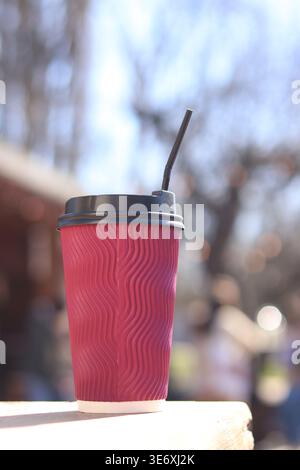 Roter Papierbecher mit gewellter Oberfläche und schwarzem Deckel und Strohhalm, der auf einem Holztisch vor einem verschwommenen Hintergrund eines Sommercafés steht. Zum Mitnehmen Stockfoto