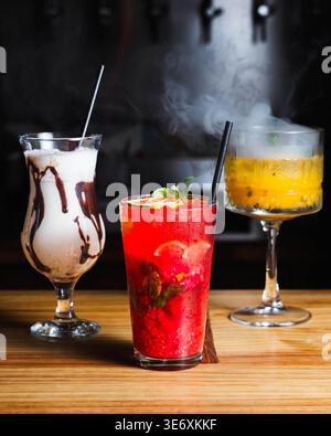 Drei bunte Cocktails ruhen auf einer Holzbar. Dampflocken aus dem tropischen Getränk in einem Weinglas. Schokoladengetränk steht neben einem leuchtenden Rot Stockfoto