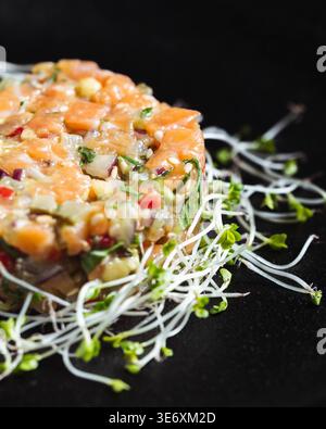 Frischer Lachs-Tatar mit knusprigen Sprossen auf schwarzem Hintergrund. Hellgrüne Triebe stehen im Kontrast zur orangen Fischstruktur. Gewürfeltes Gemüse verleiht Farbe und Fre Stockfoto