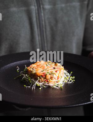 Gourmet-Fisch-Tatar sitzt auf Sprossen. Dunkle Platte steht im Kontrast zu lebendigen, frischen Zutaten. Handheld-Präsentationen weisen auf Sorgfalt und Handwerkskunst hin. S Stockfoto