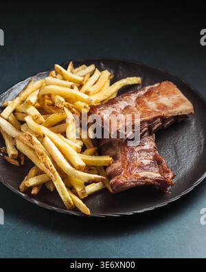 Gegrillte Rippchen sitzen neben goldenen Pommes Frites auf dunklem Teller. Textur und Farbe lassen eine zarte, aromatische Zubereitung vermuten. Einfache, auffällige Präsentation betont das Hören Stockfoto