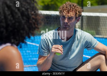 Vielseitiges Tennispaar-Training auf dem blauen Platz, Mann sitzt am Netz in Sportswear und hält Neonball Stockfoto