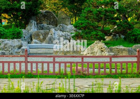 Eine ruhige Gartenszene mit einem roten Holzzaun, großen grauen Felsen und lebendigem Grün mit Bäumen und Gras, die eine friedliche Natur schaffen Stockfoto