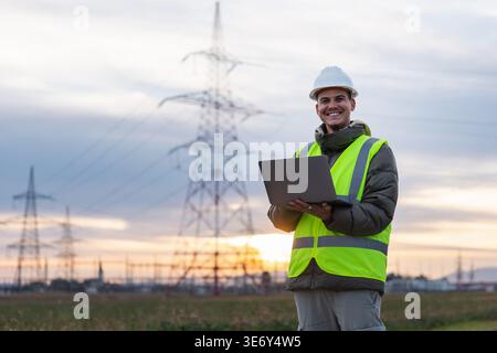 Junger männlicher Elektroingenieur Portrait führt die Inspektion von Hochspannungsleitungen mit einem Laptop im Freien während der Sonnenuntergänge durch. Stockfoto