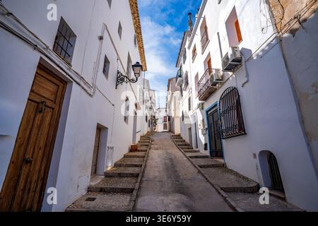 Eine malerische Kopfsteinpflasterallee gesäumt von hellen weißen mediterranen Häusern unter einem klaren blauen Himmel in Altea. Die zentrale Perspektive führt das Auge nach oben Stockfoto