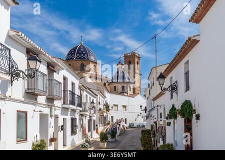 Eine malerische Gasse gesäumt von hellen weißen mediterranen Häusern in Altea, Spanien. In der Mitte stehen die legendären blau-weißen Keramikkuppeln und der Stockfoto