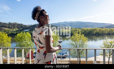 Ruhige Rückansicht einer Afrikanerin mit Wein, die von einem Balkon aus eine sonnenverwöhnte Landschaft bewundert Stockfoto