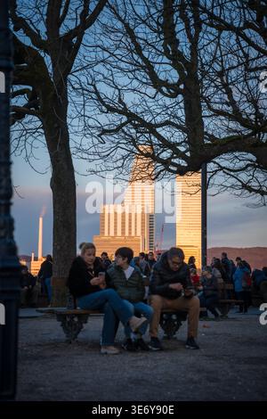 Basel, Schweiz - 10. März 2025: Blick auf Menschen, die das Abendlicht auf einer Bank genießen, mit der schroffen, geometrischen Architektur des BIZ-Turms risi Stockfoto