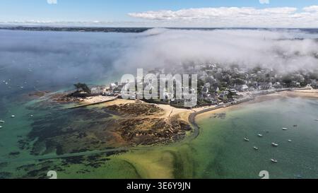 Aus der Vogelperspektive auf die nebelbelbeladene Küste trifft auf malerische Häuser und den in Nebel gehüllten Sandstrand, Carantec, Bretagne, Frankreich. Stockfoto