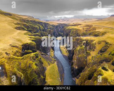 Aus der Vogelperspektive auf den Fluss, der sich durch die tiefe Schlucht schlängelt, wo moosbedeckte Klippen unter einem wilden, dramatischen Himmel auf den Himmel treffen, Fjaðrárgljúfur, Skaftá Stockfoto