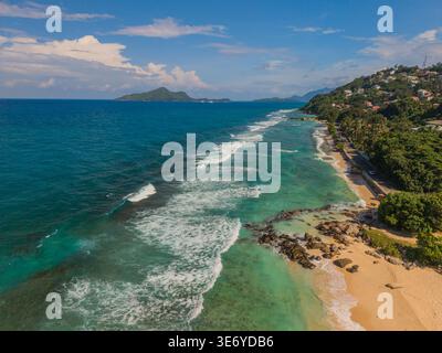 Aus der Vogelperspektive auf türkisfarbene Wellen, die sanft den goldenen Sand küssen, üppige grüne Hügel, die den azurblauen Himmel treffen, La Retraite, Mahe Island, Seychellen. Stockfoto