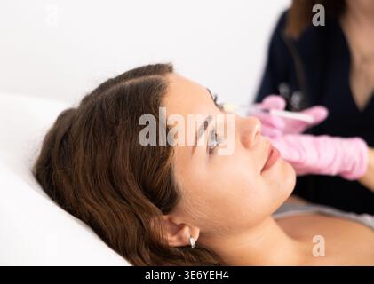 Hand des Kosmetikers in Handschuhen mit verjüngenden Injektionen Verfahren zur Straffung und Glättung von Falten auf der Gesichtshaut der jungen Frau in Schönheit Stockfoto