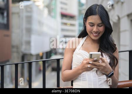 Eine lächelnde junge asiatische Frau schaut auf ihr Handy, während sie auf einem Fußweg im Freien in Central, Hongkong steht. Der Hintergrund zeigt eine verschwommene Stadt Stockfoto