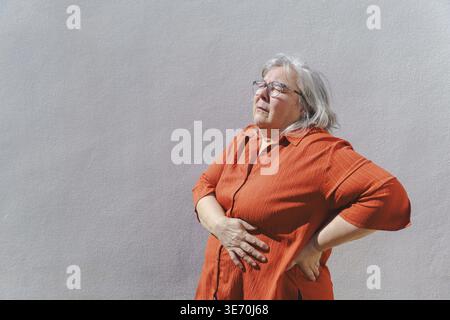 Ältere Frau leidet unter Bauchschmerzen und Rückenschmerzen, hält ihren Bauch und unteren Rücken, zeigt Beschwerden und gesundheitliche Probleme oft mit wi verbunden Stockfoto