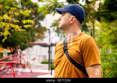 Ein Mann mit gelbem T-Shirt und blauer Baseballmütze steht im Stadtpark und blickt weg. Urbaner Lifestyle und legere Mode in grüner Outdoor-Umgebung mit Stockfoto