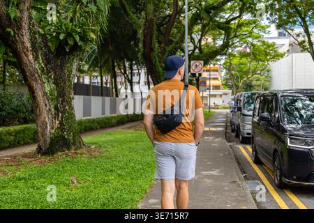 Mann in orangefarbenem T-Shirt läuft auf einem von Bäumen gesäumten Bürgersteig in Singapur. Städtischer Lebensstil und Reiseerfahrung in tropischer Stadtumgebung mit Parkplätzen ca. Stockfoto