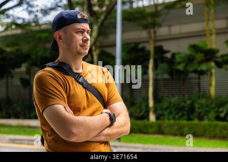Mann in gelbem T-Shirt und Baseballmütze, der mit überkreuzten Armen im Stadtpark steht. Legere männliche Reisende oder Pendler, die auf der Straße im Freien wegschauen Stockfoto