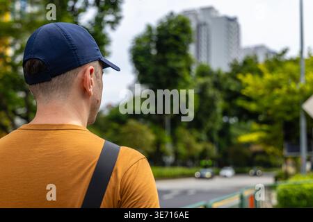 Mann mit blauer Mütze und orangefarbenem T-Shirt, der auf die Stadtstraße mit grünen Bäumen und Gebäuden blickt. Rückansicht der männlichen Touristen, die Metropolitan erkunden Stockfoto