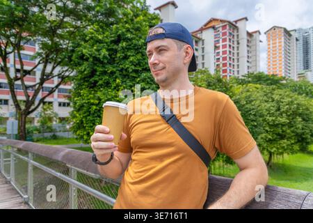Mann mit Kaffeetasse zum Mitnehmen auf einer hölzernen Parkbrücke in Singapur mit HDB-Wohngebäuden im Hintergrund, aufgenommen als Taille-up-Porträt. Stockfoto