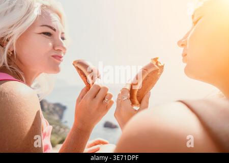 Freunde, die im Freien Gebäck essen, genießen einen Sommer-Snack an einem malerischen Blick auf die Küste bei hellem Tageslicht Stockfoto