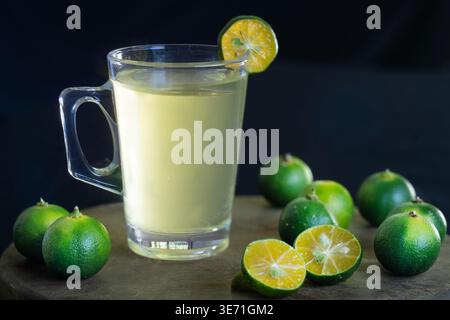 Ein erfrischendes Glas Calamansi-Saft, serviert in einem Becher mit einer Scheibe Beilage, umgeben von frischen Calamondin-Früchten und grünen Blättern Stockfoto