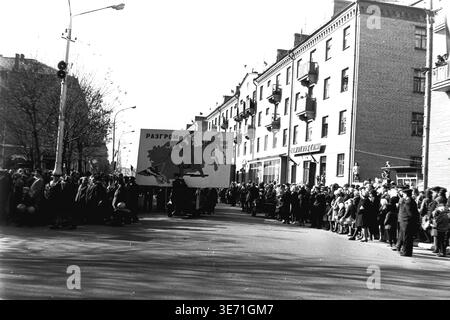 Slaviansk, 1960er Jahre, Platz der Oktoberrevolution (heute Soborna-Platz). Eine riesige Menschenmenge versammelt sich entlang der Straße, um eine feierliche sowjetische Demonstration zu sehen. Im Mittelpunkt steht eine große mobile Propaganda-Werbetafel, die durch die Prozession getragen wird und eine eindrucksvolle Illustration eines sowjetischen Soldaten darstellt, der ein faschistisches Hakenkreuz über einer Karte zerstört. Der kühne russische Text lautet: "Niederlage des faschistischen Deutschlands". Zuschauer säumen die Gehwege in der Nähe von Wohngebäuden. Friedliches Leben im Donbass vor dem Konflikt. Stockfoto