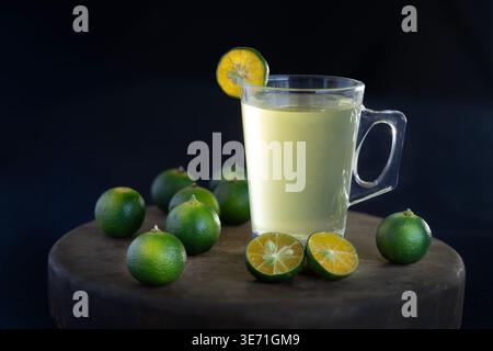 Ein erfrischendes Glas Calamansi-Saft, serviert in einem Becher mit Stielen und einer Scheibe Beilage, umgeben von frischen Calamondin-Früchten und grünen Blättern Stockfoto