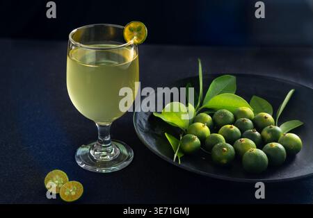 Ein erfrischendes Glas Calamansi-Saft, serviert in einem Becher mit Stielen und einer Scheibe Beilage, umgeben von frischen Calamondin-Früchten und grünen Blättern Stockfoto