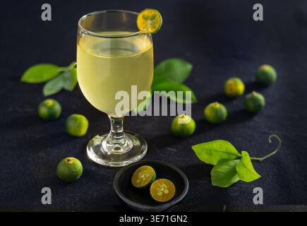Ein erfrischendes Glas Calamansi-Saft, serviert in einem Becher mit Stielen und einer Scheibe Beilage, umgeben von frischen Calamondin-Früchten und grünen Blättern Stockfoto