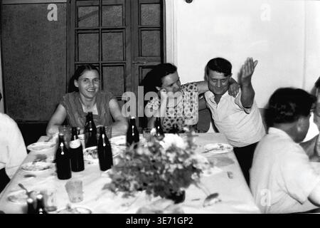 Slaviansk, 1959, Energy Technical College. Drei glückliche Teilnehmer werden am festlichen Banketttisch festlich festlich festlich festlich festlich festlich festlich festlich festlich Ein Mann auf der rechten Seite hebt enthusiastisch die Hand in einer fröhlichen, animierten Geste und sitzt liebevoll mit dem Arm um eine lachende Frau. Eine andere Frau sitzt ruhig links. Der Tisch ist mit einem hübschen floralen Herzstück dekoriert und mit Tellern, Gläsern und dunklen Getränkeflaschen verstreut. Friedliches Leben im Donbass vor dem Konflikt. Stockfoto