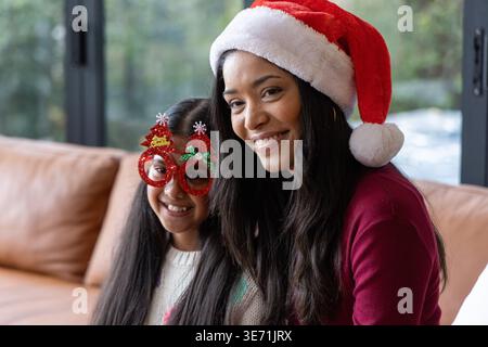Mutter und Tochter sitzen im Wohnzimmer auf einer braunen Ledercouch mit Weihnachtsmütze und Glitzerbrille Stockfoto