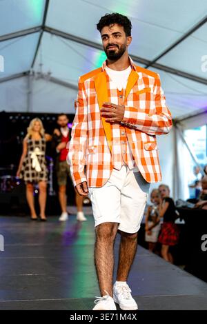 Villach, Österreich - 18. Juli 2025: Männliches Model mit orangefarbenem kariertem Blazer und weißen Shorts geht auf einer Modeschau mit zusätzlichem Mo über den Laufsteg Stockfoto