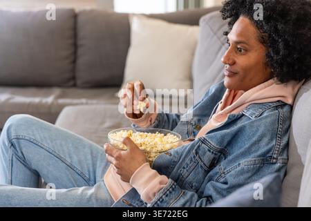 Afroamerikanerin, die sich zu Hause auf dem Sofa in Denim liegend, Glasschale Popcorn, Kopierraum. Gemütlich, Zuhause, Entspannung, lässig, weich, natürlich, Neutr Stockfoto