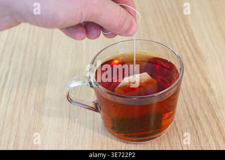 Teebeutel mit Hand in eine transparente Glasschale mit heißem Tee auf Holztisch, einfacher Getränkezubereitung und gemütlichem Hausgetränk-Konzept Stockfoto