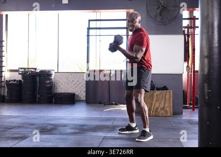 Afroamerikaner mit Hexenhantel, Bizepscurl im Fitnessstudio in der Nähe von Tellern, Kopierraum. Athlet, Gewichte, Training, Kraft, Sonnenlicht, einstauben Stockfoto