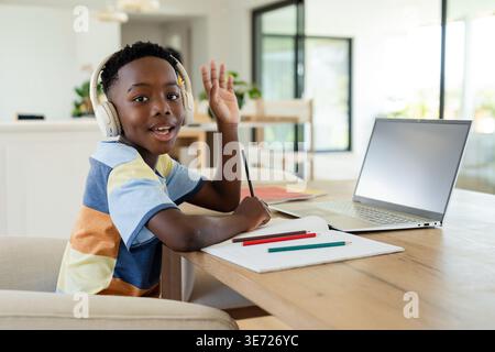 Afroamerikanisches Kind sitzt am Tisch in gestreiftem T-Stück, Headset, Schreiben mit Farbstiften, Laptop. Student, zu Hause, studieren, Fokus, Notizbuch, Kopfhörer Stockfoto