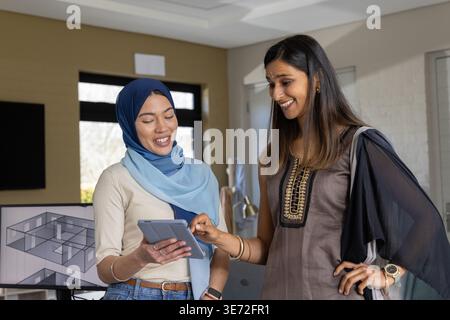 Verschiedene weibliche Kollegen in Hijab und Kurta, die Tablets überprüfen und im Büro auf den Plan zeigen. Zusammenarbeit, Interior, modern, Design, Layout, Stockfoto
