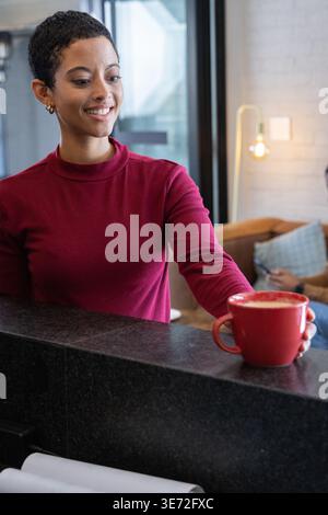 Afroamerikanerin, die an der Cafeteke arbeitet, legt rote Tasse mit Schaum und trägt einen burgunderroten Pullover. Barista, Kaffeehaus, Latte, Keramik, Tasse, Tageslicht Stockfoto