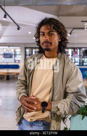 Indischer Mann steht im Café, lehnt sich auf einen Pflanztopf, trägt Bomberjacke und schwarze Smartwatch. Modern, Coworking, Interior, Green, Industrial, Colla Stockfoto