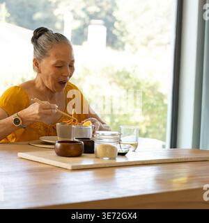 Ältere asiatische Frau in Senfbluse, die zu Hause mit Stäbchen aus Schüssel, Kopierraum isst. Ältere Menschen, Frauen, Essen, Nudeln, Utensilien, Sonnenlicht, g Stockfoto
