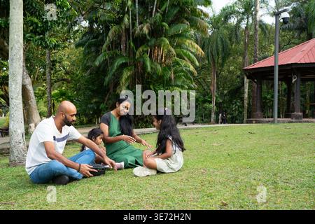 Indische Familie, die auf grünem Gras sitzt, sich in einem üppigen Park entspannt und entspannt Stockfoto