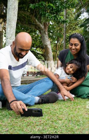 Indische Familie, die einen glücklichen Moment mit dem Smartphone in einem üppigen grünen Park festnimmt Stockfoto