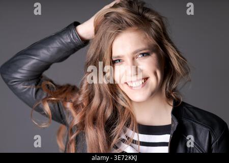 Eine Frau mit langen braunen Haaren lächelt und posiert für ein Foto. Sie trägt eine schwarze Jacke und ein weißes gestreiftes Hemd Stockfoto