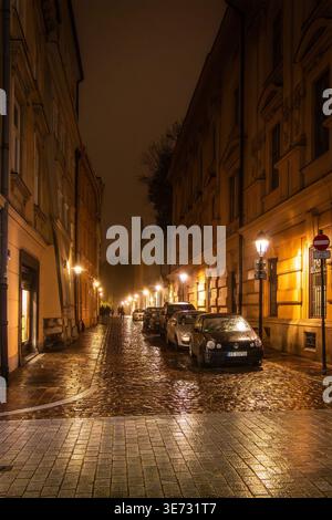 Nächtlicher Blick auf eine enge Kopfsteinpflasterstraße in Krakau Polen mit geparkten Autos und beleuchteten historischen Gebäuden unter dunklem Himmel Stockfoto