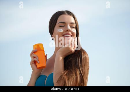 Eine schöne junge Frau lächelt, als sie am Strand Sonnencreme auf ihr Gesicht aufträgt. Die Sonne scheint hell und unterstreicht ihr Engagement für die Hautpflege Stockfoto