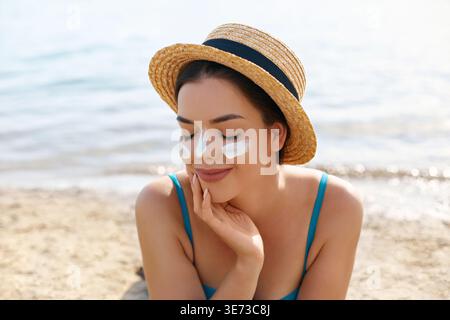 Eine junge Frau mit Strohhut sitzt am Sandstrand und lächelt vor Freude, während sie Sonnencreme auf ihr Gesicht aufträgt. Die Sonne scheint hell über den ruhigen s Stockfoto