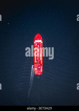Aus der Vogelperspektive eines leuchtenden roten Bootes, das durch das dunkle, strukturierte Wasser vor der Küste, Ballstad, Nordland, Norwegen, schneidet. Stockfoto