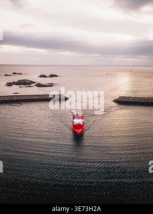 Aus der Vogelperspektive eines leuchtenden roten Schiffes, das durch das ruhige, dunkle Wasser in Richtung Horizont fährt, eingerahmt von Steinbrechern, Ballstad, Nordland, Norwa Stockfoto