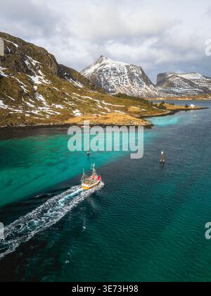 Aus der Vogelperspektive eines Fischerbootes, das durch türkisfarbenes Wasser schneidet, im Kontrast zu den zerklüfteten, schneebedeckten Bergen von Kåkernbrua, Ramberg, Nordland, N Stockfoto