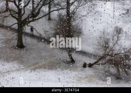 Regentropfen auf Fensterglas mit verschwommenem Winterparkhintergrund, melancholische Atmosphäre Stockfoto